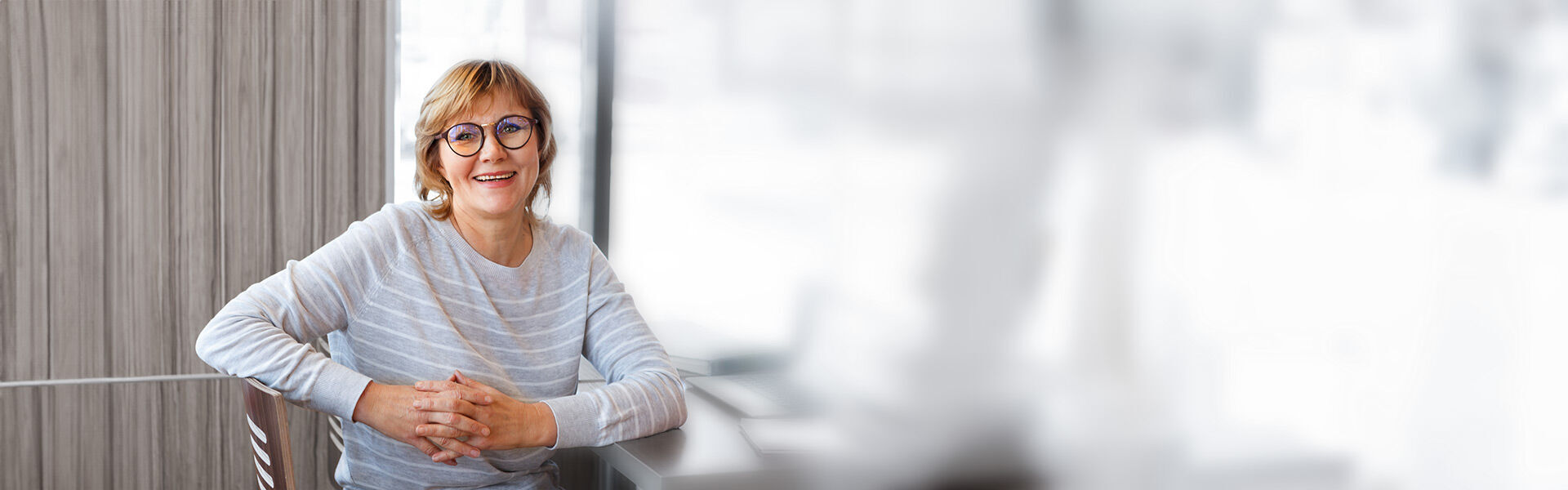 Frau mittleren Alters sitzt entspannt auf einem Stuhl und blickt freundlich in die Kamera.