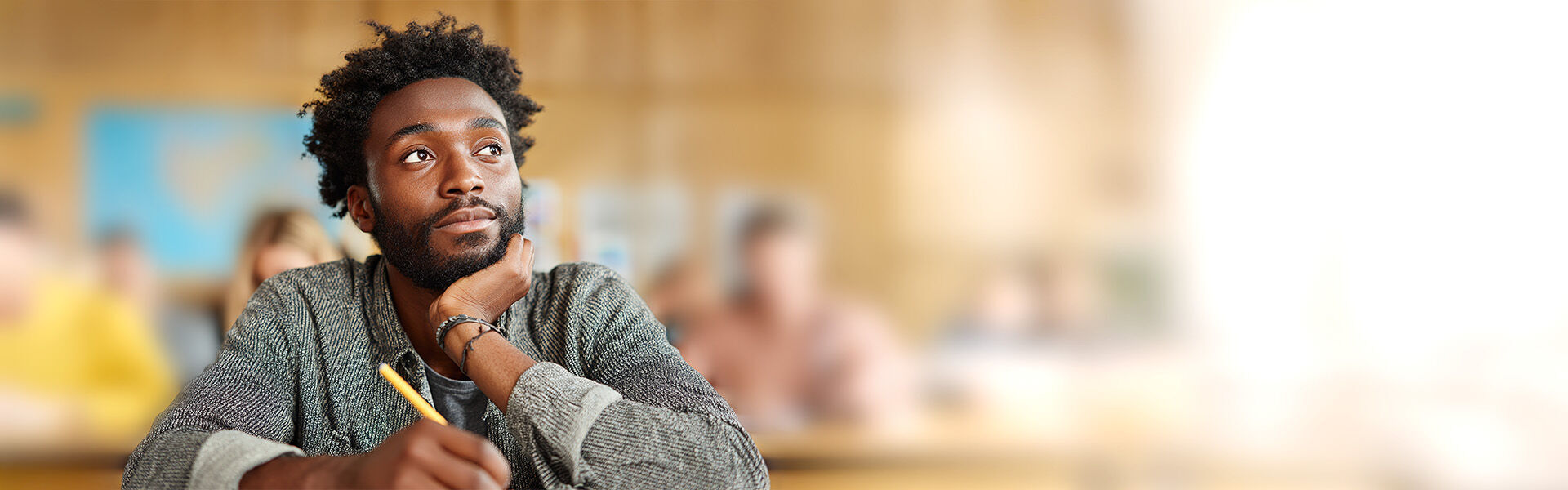 Ein dunkelhäutiger Mann sitzt im Klassenzimmer mit einem Stift in der Hand, das Kinn in der anderen Hand, und schaut nachdenklich zur Seite, während er über seiner Aufgabe brütet.
